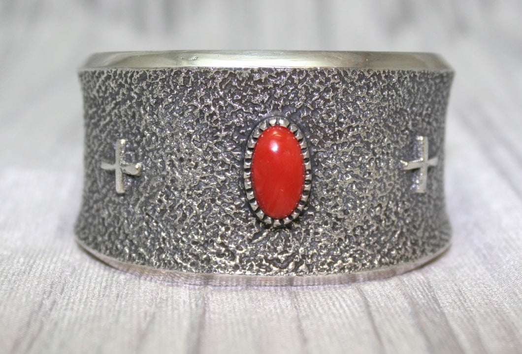 A wide silver cuff bracelet with a textured surface, featuring a bright oval red stone at the center and two small silver crosses on each side. The cuff rests on a soft gray background, highlighting its intricate craftsmanship.