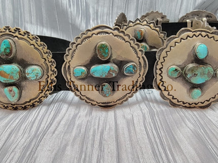 A handcrafted Navajo concho belt with detailed sterling silver conchos, each embedded with mixed turquoise stones of varying shades and natural patterns, attached to a black leather strap.