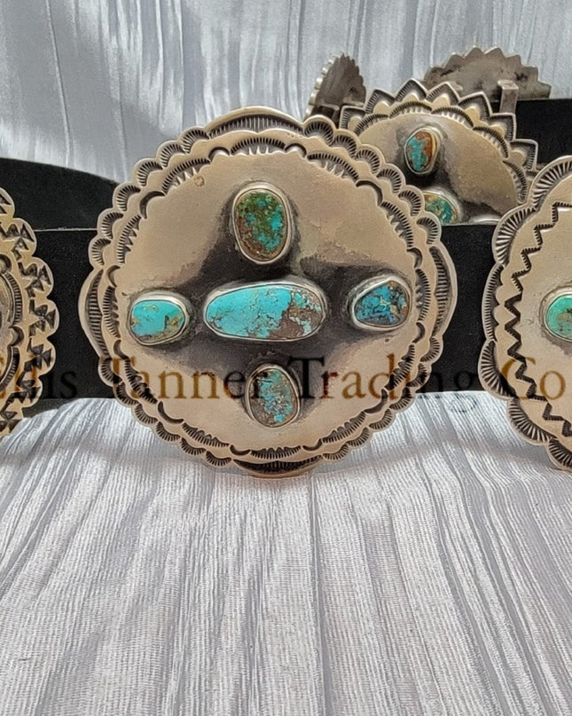 A handcrafted Navajo concho belt with detailed sterling silver conchos, each embedded with mixed turquoise stones of varying shades and natural patterns, attached to a black leather strap.
