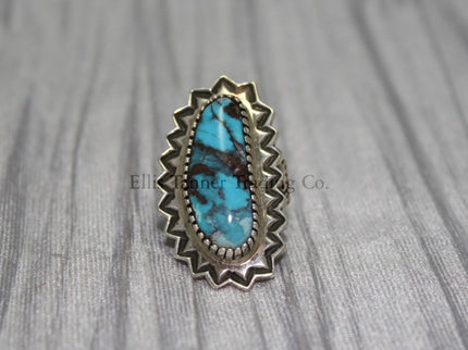 An oval-shaped Bisbee Turquoise with black vein matrix. surrounded in a silver starburst border. this ring is displayed on top of a gray cloth 
