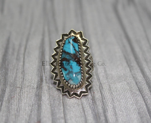 An oval-shaped Bisbee Turquoise with black vein matrix. surrounded in a silver starburst border. this ring is displayed on top of a gray cloth 