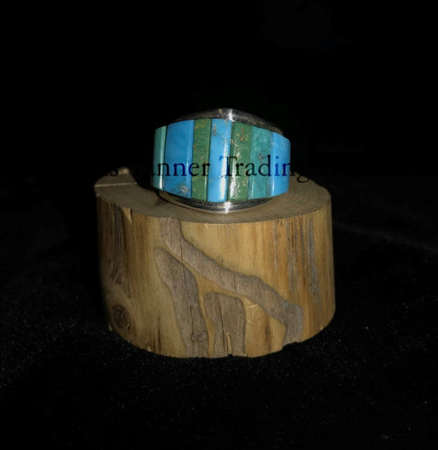 A silver-banded ring with blue and green stone inlays, displayed on a wooden piece, with the brand name 'Inner Trading' visible.