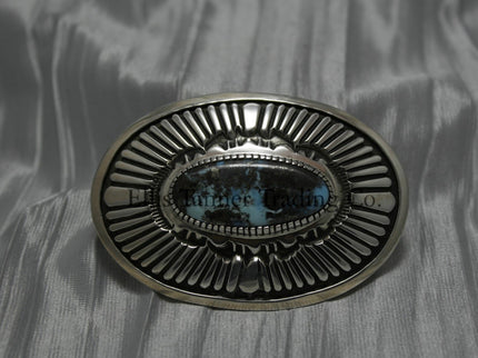 Oval shaped buckle, with a center Black diamond turquoise stone, displayed on top of a shiny silver sheet 