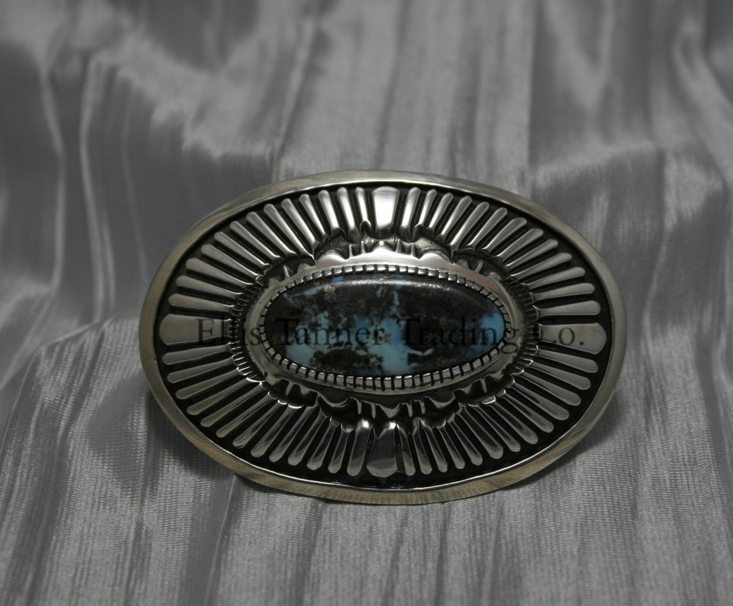 Oval shaped buckle, with a center Black diamond turquoise stone, displayed on top of a shiny silver sheet 