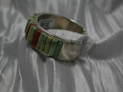 A silver bracelet featuring an intricate cobblestone inlay design with vibrant Carico Lake turquoise and coral stones. The piece is displayed on a soft, textured silver-gray fabric, side profile 