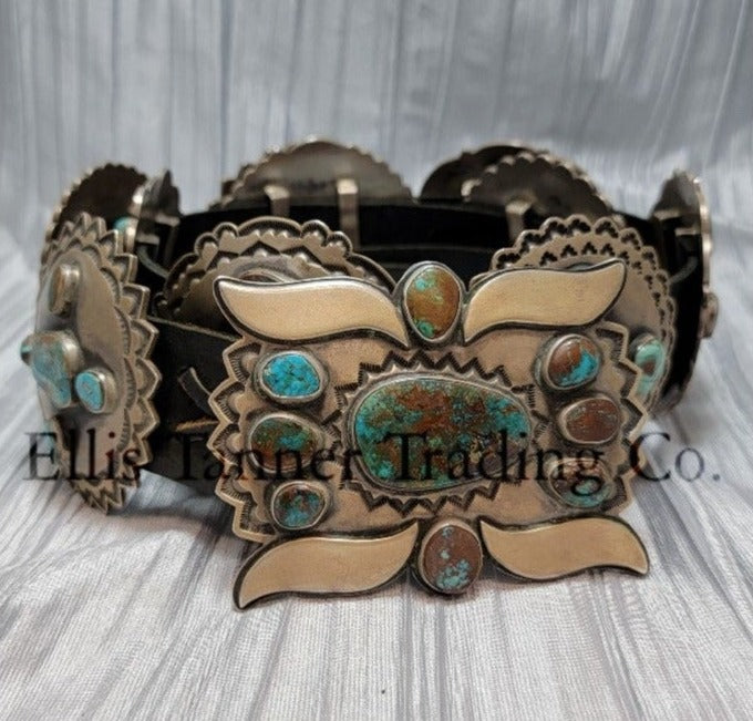 A handcrafted Navajo concho belt with detailed sterling silver conchos, each embedded with mixed turquoise stones of varying shades and natural patterns, attached to a black leather strap. Buckle visible 