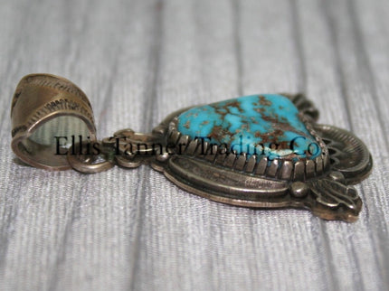 Silver Pendant, over lay silver with hand stamped designs. A center Kingman stone surrounded by a silver bezel. placed on top of a shiny cloth 