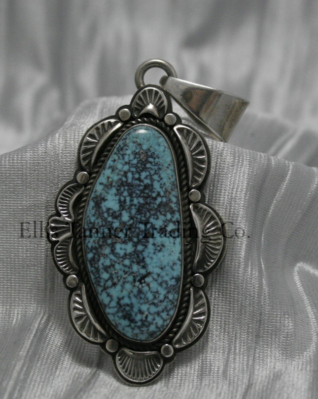 A handcrafted sterling silver pendant with a Morenci Turquoise gemstone. The stone is blue with a black and gray matrix and is set in an ornate silver frame with scalloped and stamped detailing. The pendant features a large bail for easy attachment to a chain.