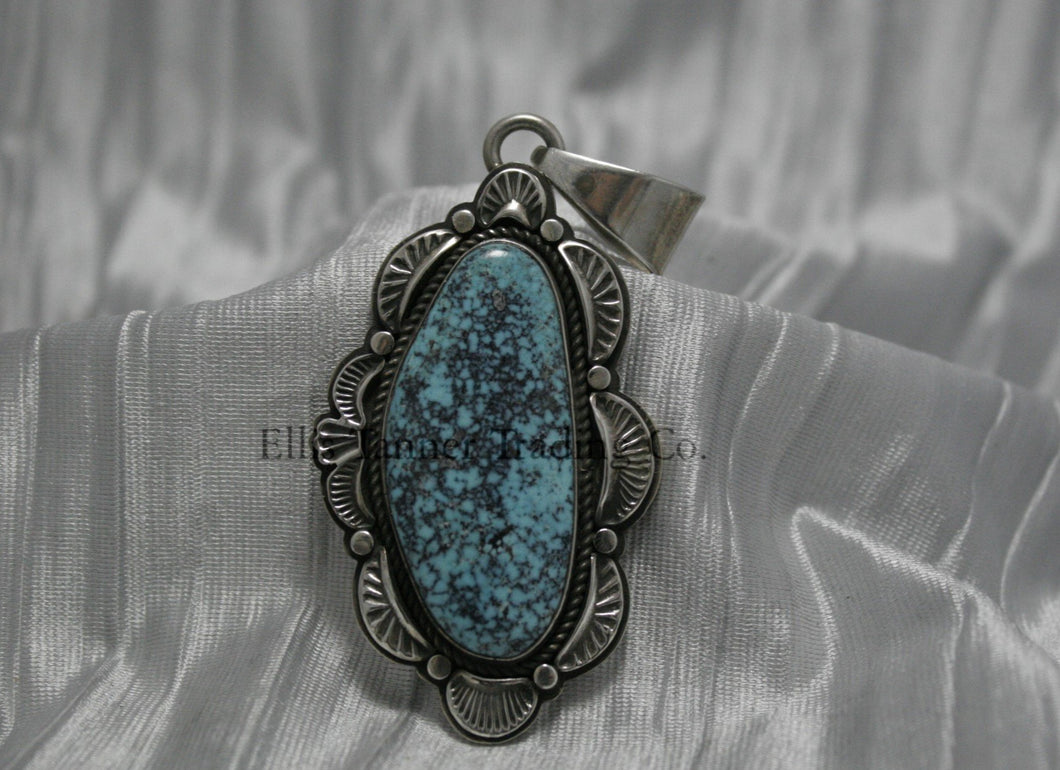 A handcrafted sterling silver pendant with a Morenci Turquoise gemstone. The stone is blue with a black and gray matrix and is set in an ornate silver frame with scalloped and stamped detailing. The pendant features a large bail for easy attachment to a chain.