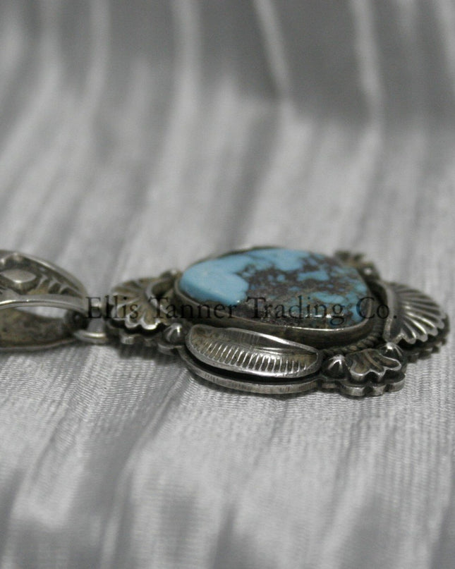 Teardrop-shaped Bisbee turquoise pendant set in sterling silver with intricate engraved details, scalloped accents, and an ornate bail, displayed on a white satin background.
