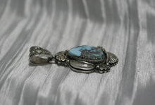 Load image into Gallery viewer, Teardrop-shaped Bisbee turquoise pendant set in sterling silver with intricate engraved details, scalloped accents, and an ornate bail, displayed on a white satin background.

