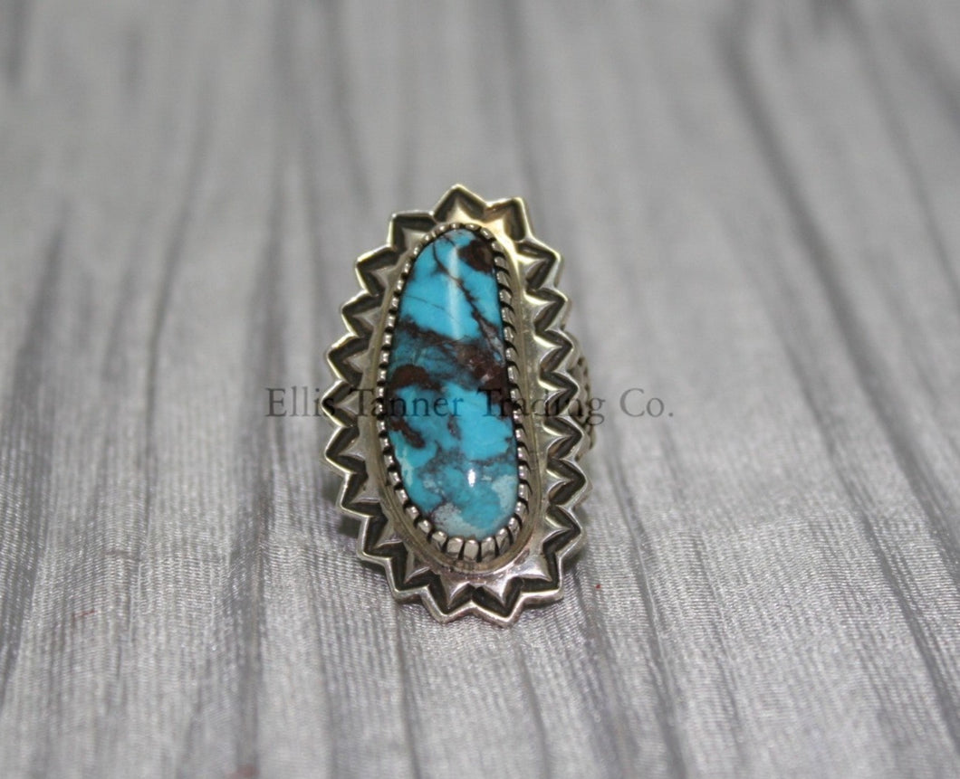 An oval-shaped Bisbee Turquoise with black vein matrix. surrounded in a silver starburst border. this ring is displayed on top of a gray cloth 