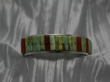 A silver bracelet featuring an intricate cobblestone inlay design with vibrant Carico Lake turquoise and coral stones. The piece is displayed on a soft, textured silver-gray fabric.