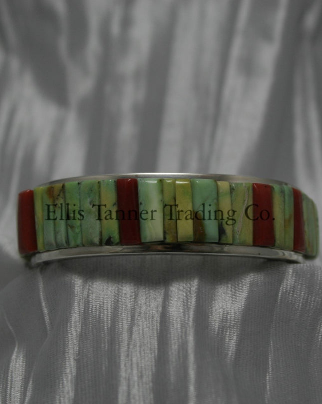 A silver bracelet featuring an intricate cobblestone inlay design with vibrant Carico Lake turquoise and coral stones. The piece is displayed on a soft, textured silver-gray fabric.
