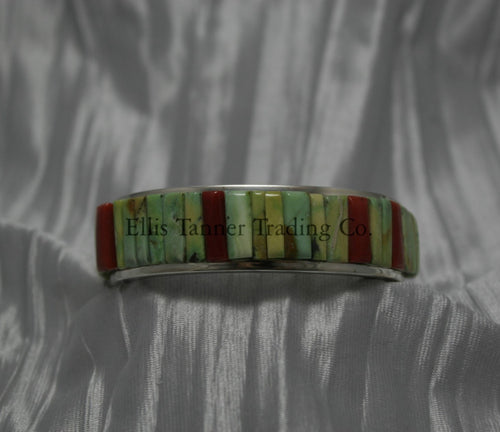 A silver bracelet featuring an intricate cobblestone inlay design with vibrant Carico Lake turquoise and coral stones. The piece is displayed on a soft, textured silver-gray fabric.
