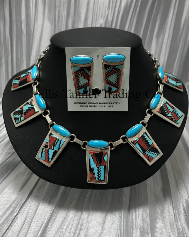 A Southwest Inlayed necklace and matching earrings set with sterling silver Inlayed with vibrant turquoise, coral, and Onyx geometric patterns. The necklace has multiple rectangular pendants, each with turquoise stones, and the earrings match the necklace design. Displayed on a black stand.