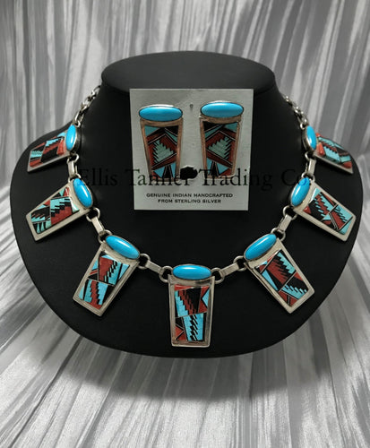 A Southwest Inlayed necklace and matching earrings set with sterling silver Inlayed with vibrant turquoise, coral, and Onyx geometric patterns. The necklace has multiple rectangular pendants, each with turquoise stones, and the earrings match the necklace design. Displayed on a black stand.