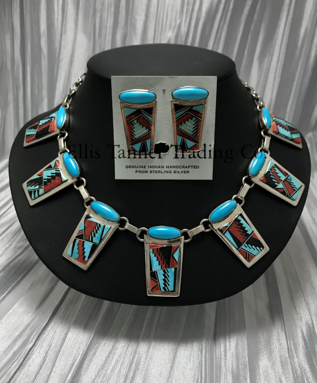 A Southwest Inlayed necklace and matching earrings set with sterling silver Inlayed with vibrant turquoise, coral, and Onyx geometric patterns. The necklace has multiple rectangular pendants, each with turquoise stones, and the earrings match the necklace design. Displayed on a black stand.