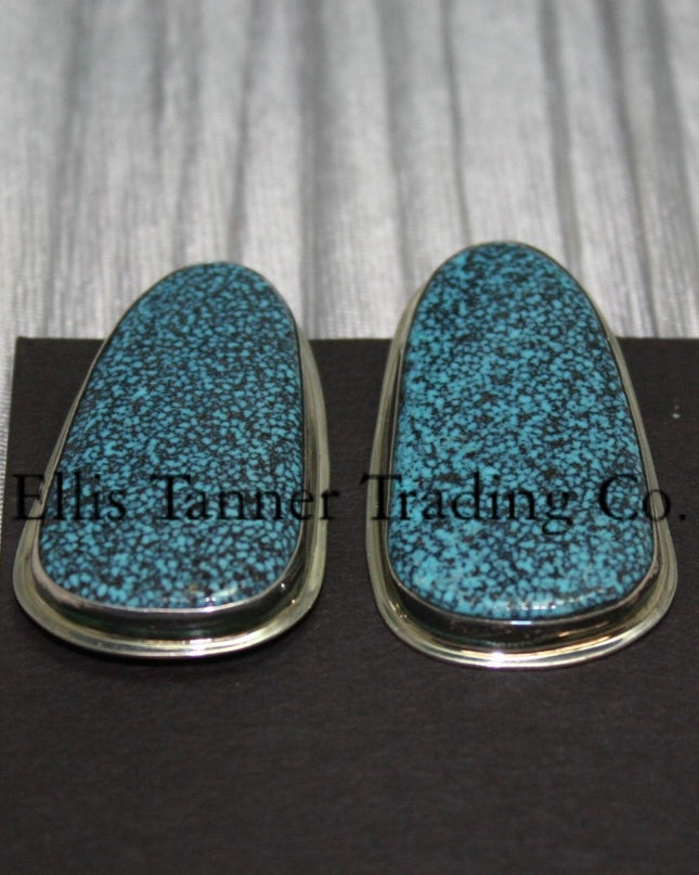 slab-style clip-on earrings featuring polished #8 turquoise stones with a speckled pattern, set in sterling silver bezels, displayed on a black card against a textured silver background. 