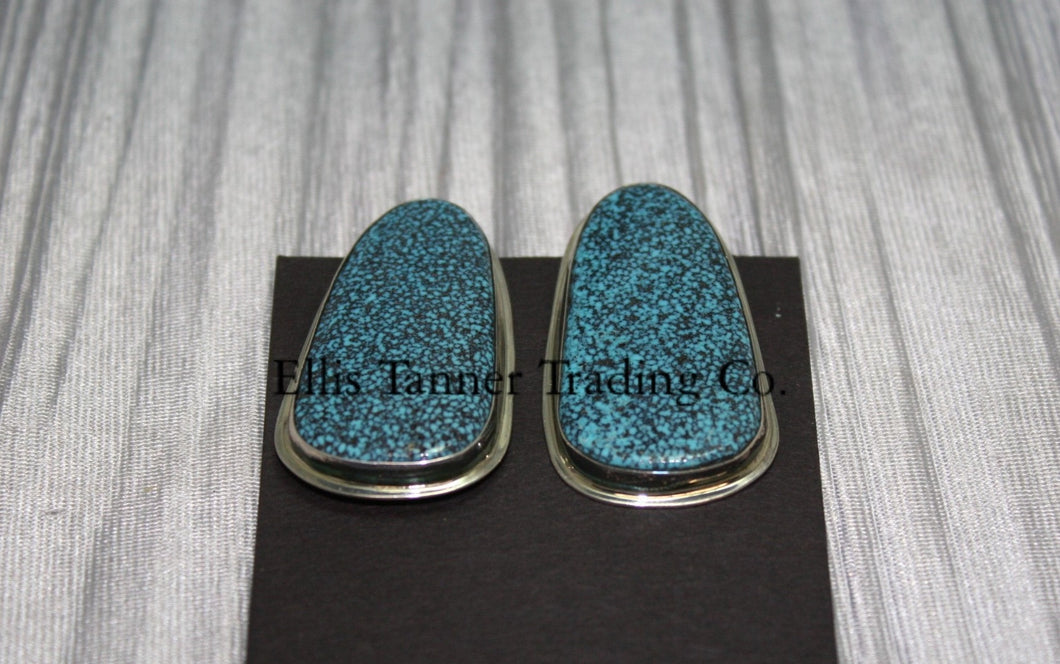 slab-style clip-on earrings featuring polished #8 turquoise stones with a speckled pattern, set in sterling silver bezels, displayed on a black card against a textured silver background. 