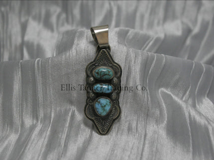 A hand stamped silver pendant with three turquoise stones arranged vertically, each set in a bezel. The pendant features intricate geometric and linear stamped patterns on the silver, It has a sturdy silver bail at the top and an elongated, curved shape.
