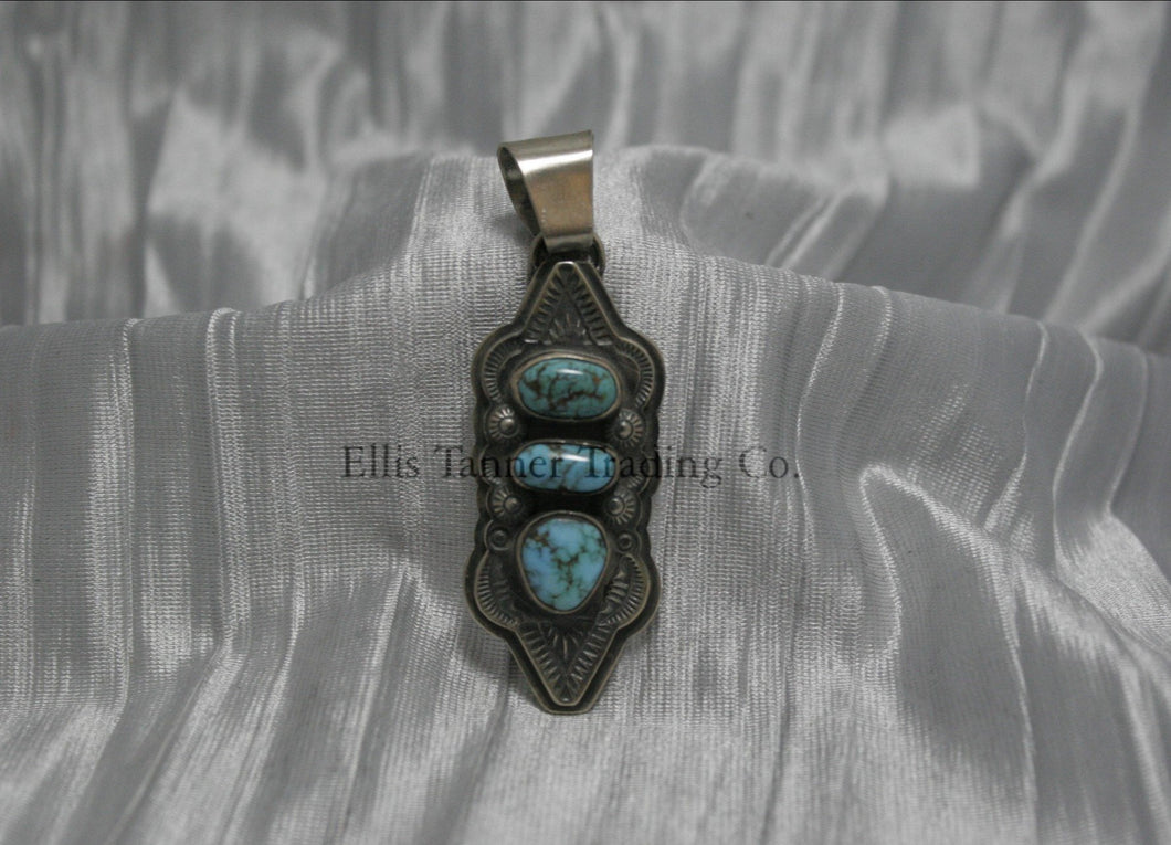 A hand stamped silver pendant with three turquoise stones arranged vertically, each set in a bezel. The pendant features intricate geometric and linear stamped patterns on the silver, It has a sturdy silver bail at the top and an elongated, curved shape.