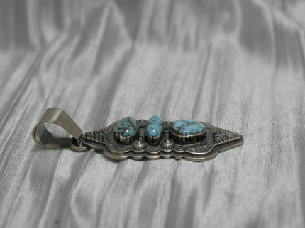 A hand stamped silver pendant with three turquoise stones arranged vertically, each set in a bezel. The pendant features intricate geometric and linear stamped patterns on the silver, It has a sturdy silver bail at the top and an elongated, curved shape.Side View 
